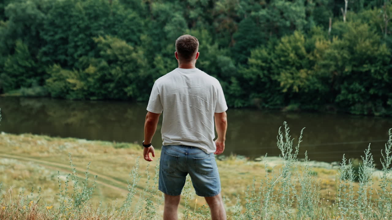 Rear view of a sportive muscular man in white t-shirt and jeans shorts. Happy person in the nature surrounding enjoying life.