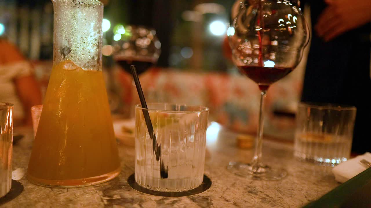 A hand pours red wine into a glass on a dimly lit table with drinks and a warm ambiance