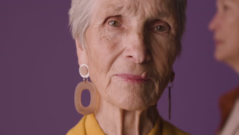 vista de cerca de una anciana rubia con el pelo corto usando camisa y chaqueta de color mostaza y aretes, posando con una mujer madura borrosa sobre fondo morado 1