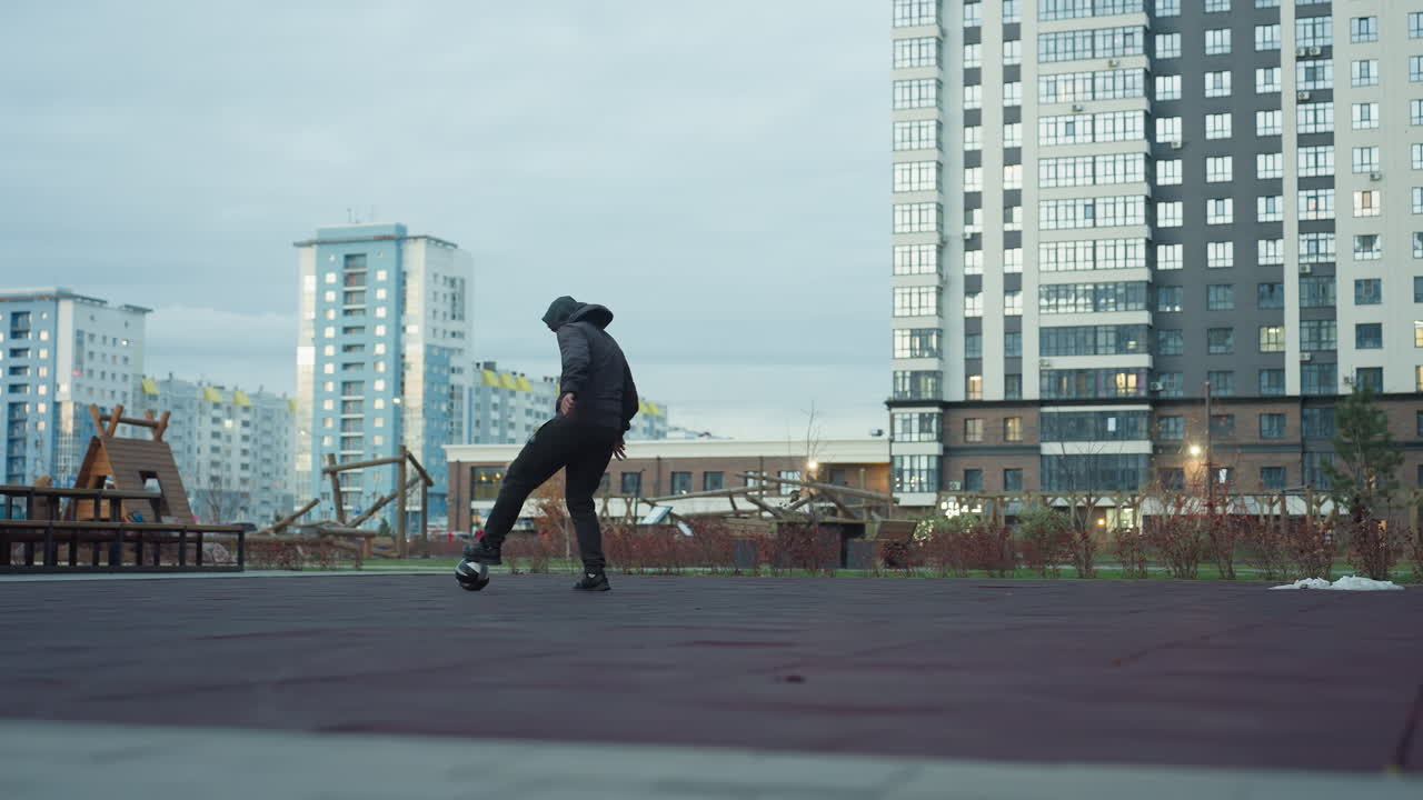 Man skillfully juggling football on sport arena during practice session, background includes urban buildings, playground, greenery, and outdoor elements