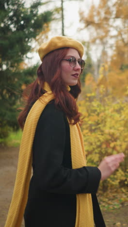 chica elegante con boina amarilla y silenciador sonríe cálidamente, tocando su cabello ondulado rojo mientras camina por el parque observando su entorno mientras pasea en un jardín vibrante