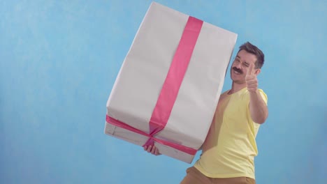 un hombre feliz y gracioso con bigote sosteniendo un gran regalo envuelto de pie sobre un fondo azul