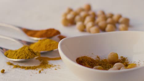 Chickpeas-fall-in-slow-motion-into-a-white-bowl-filled-with-curry-powder,-other-chickpeas-and-three-spoons-with-curry-powder,-saffron-and-turmeric-powder-are-also-seen-on-the-table