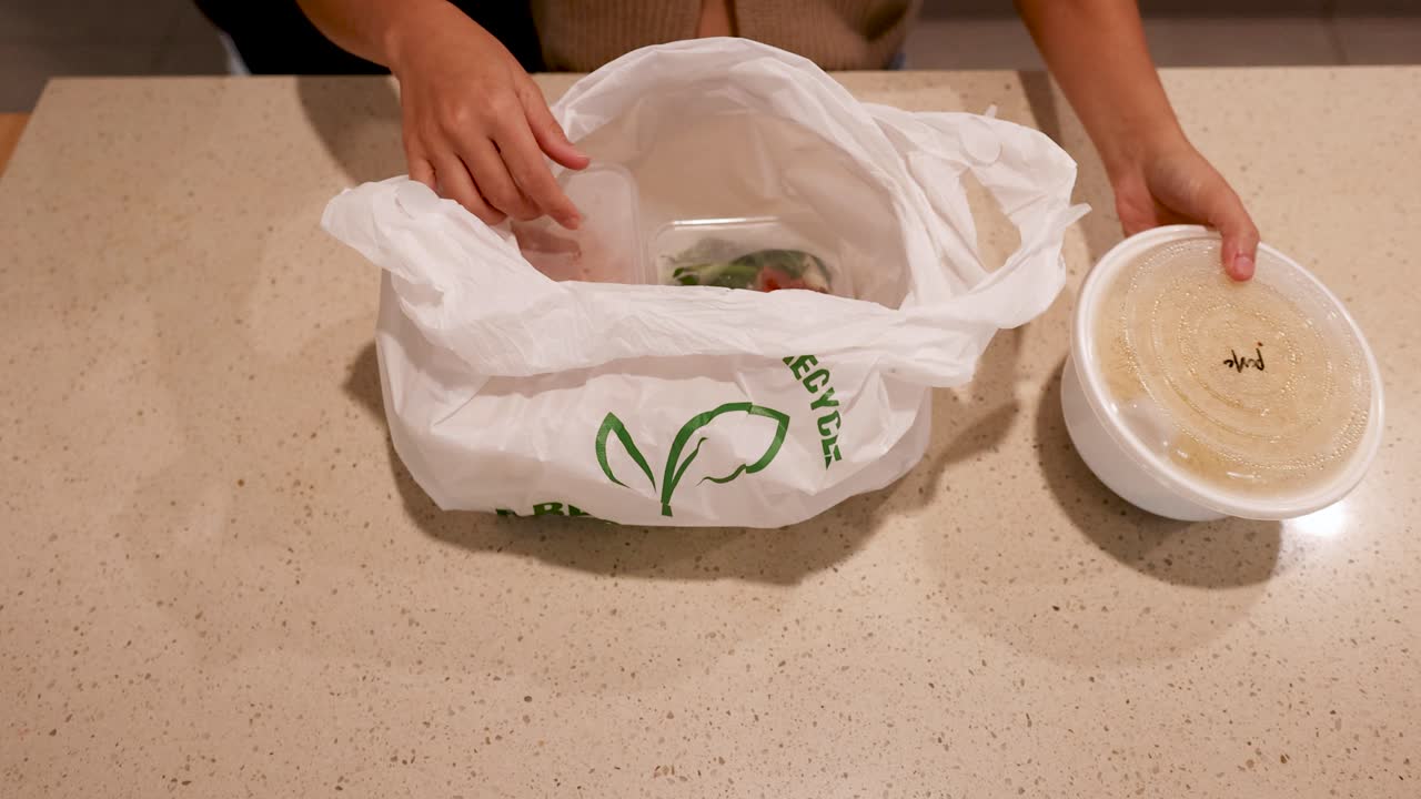 Hands unpacking Asian food from recyclable bags on a kitchen counter under warm lighting