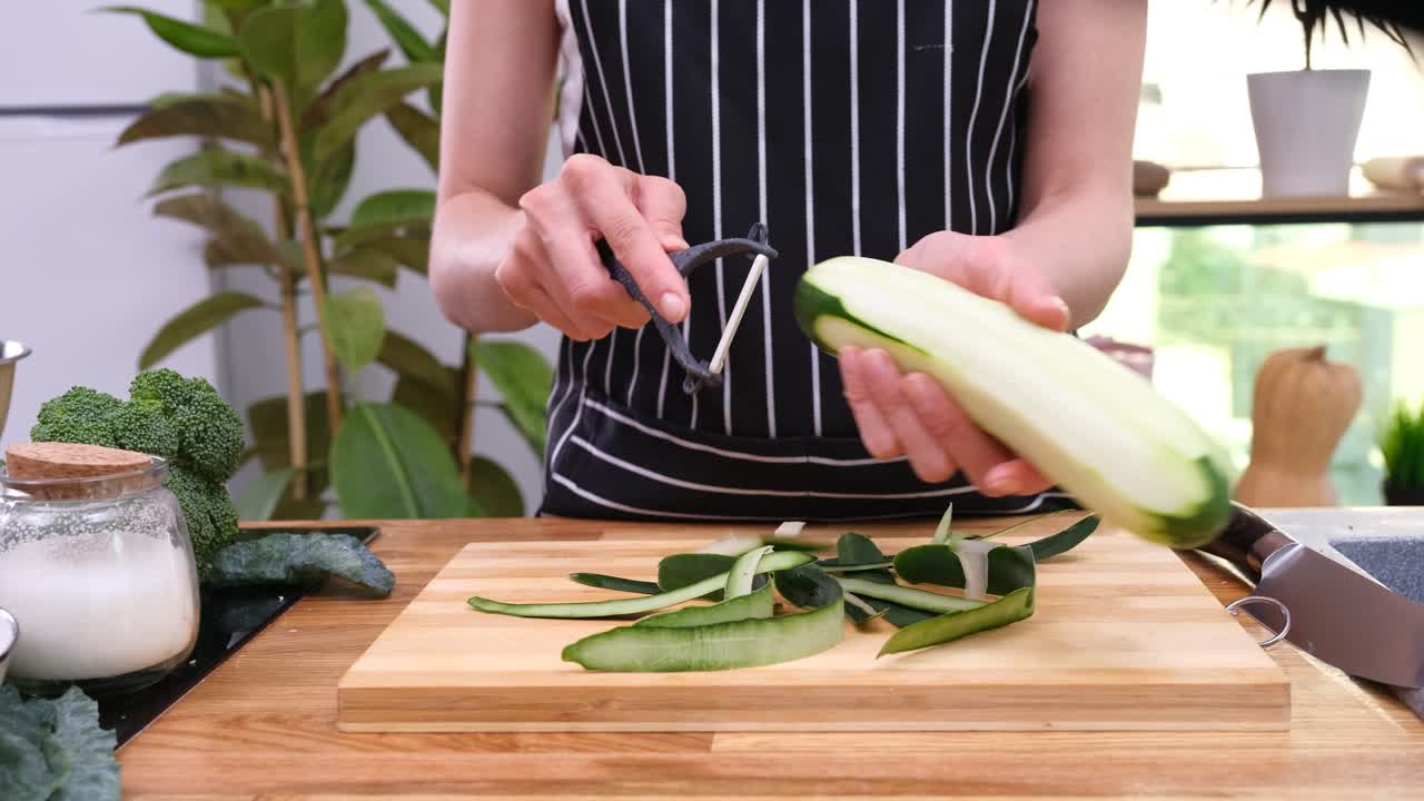 Una persona sbuccia delle zucchine impilando le strisce su un tagliere di legno.