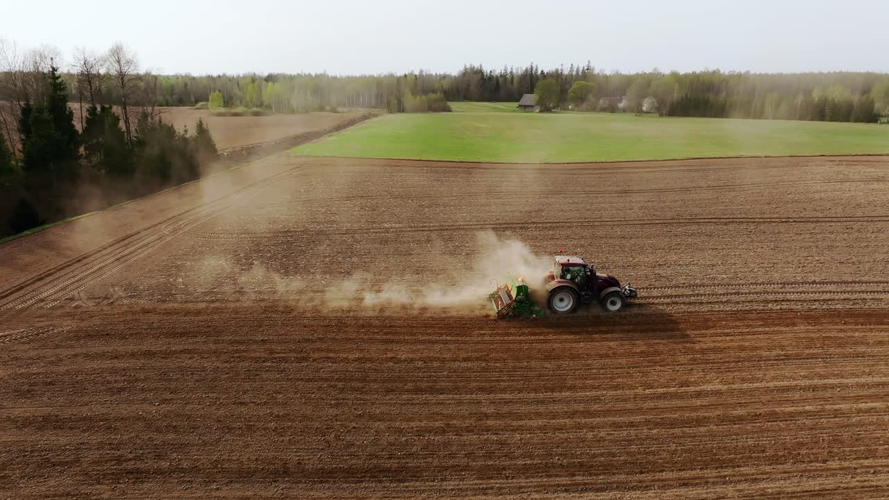 Spring tractor plows dry soil as drone flies leftward in dusty Latvian field