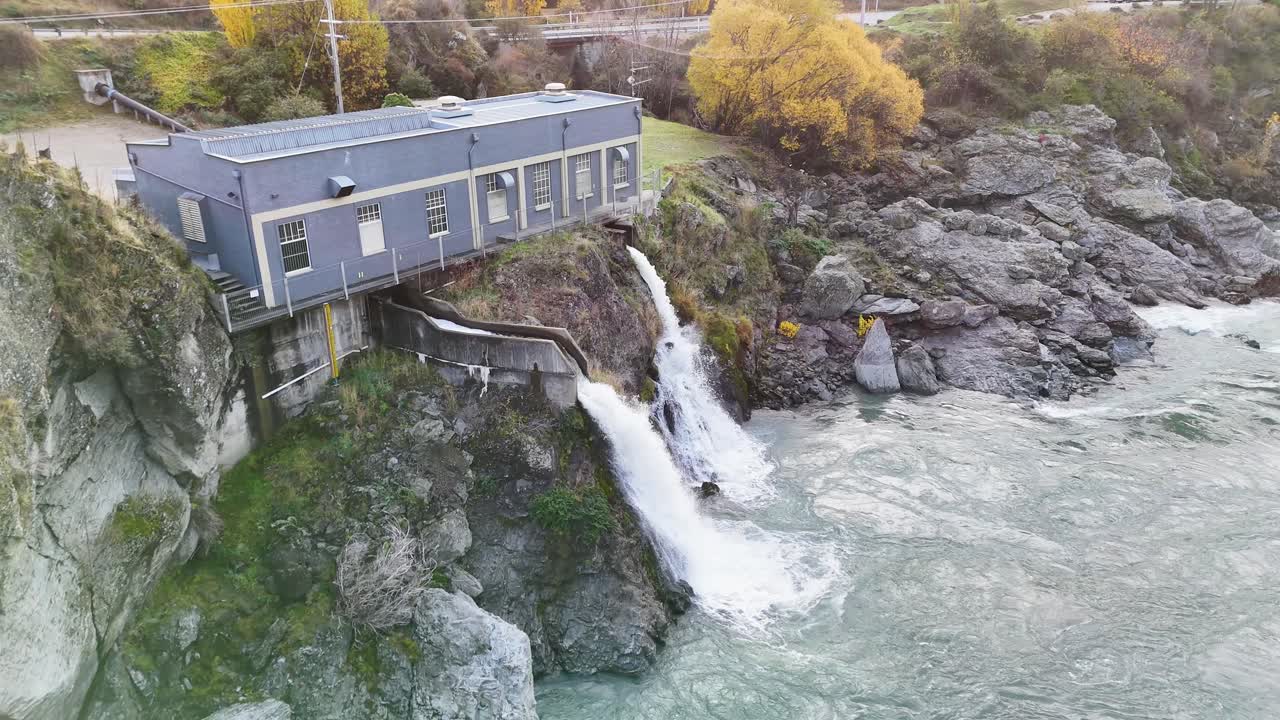 Drone footage captures a hydroelectric power station by a river, surrounded by autumn foliage and rocky terrain