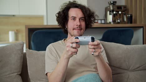 Happy-brunette-guy-with-curly-hair-and-mustache-in-a-gray-T-shirt-plays-on-a-white-joystick-and-sits-on-the-sofa-during-his-time-on-the-weekend