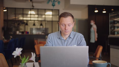 a young man in a shirt is sitting at a table with a laptop and typing on the keyboard. a student can study remotely. a businessman conducts his business remotely