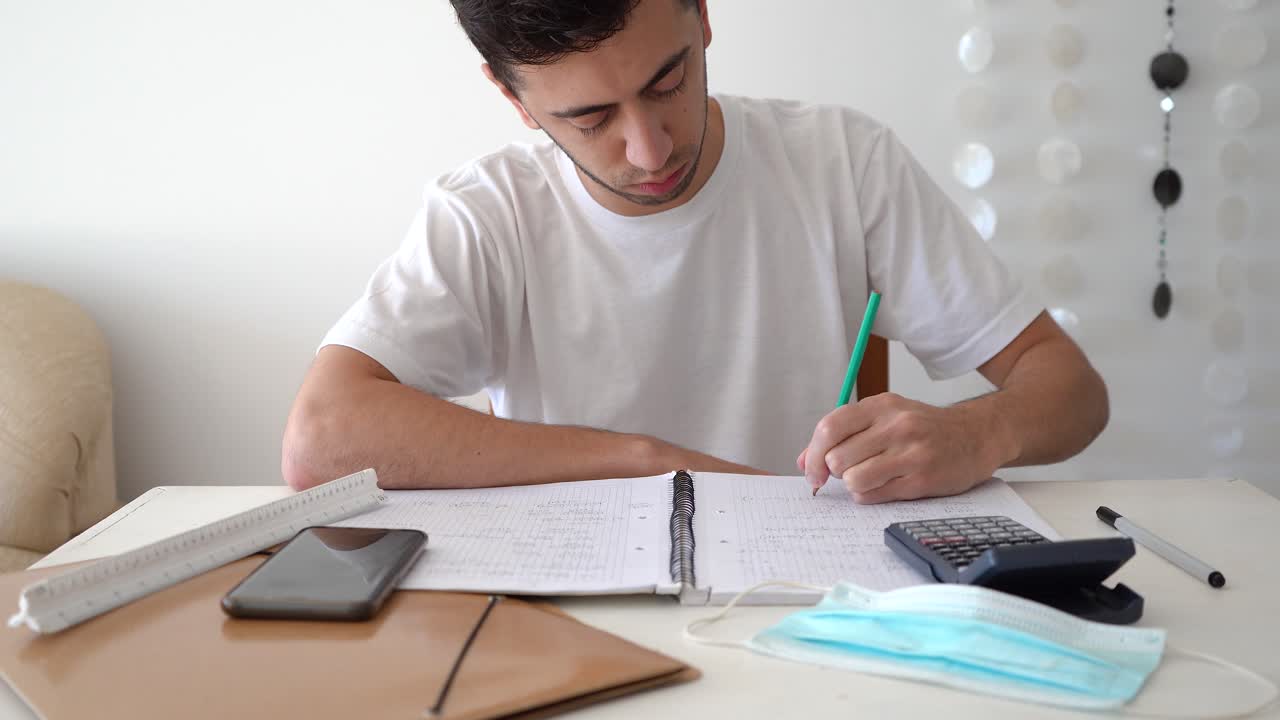 estudiante masculino escribiendo fórmulas matemáticas y soluciones en un cuaderno con calculadora, teléfono inteligente y máscara médica en la mesa