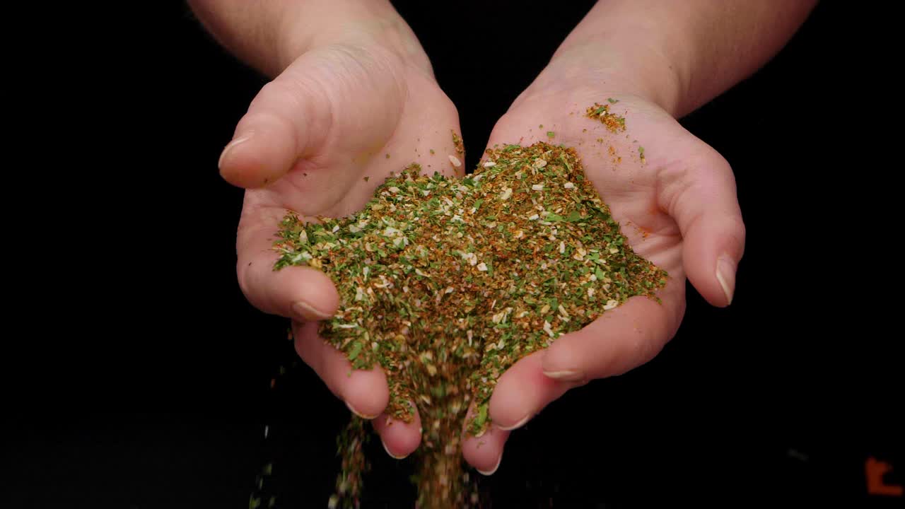 바질, 후추, 파슬리를 곁들인 맛있는 향신료를 검은 배경에 슬로우 모션으로 떨어뜨리는 여성 손의 정적인 샷