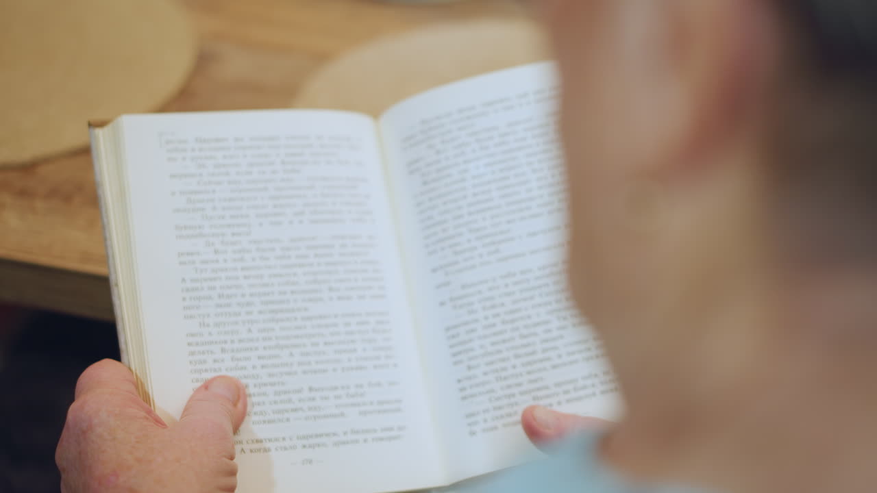Grandma reading book hands holding pages, warm kitchen table, soft natural light, vintage paperback, focused slowpaced moment, quiet intimacy, domestic setting, textured paper, thumb turning page