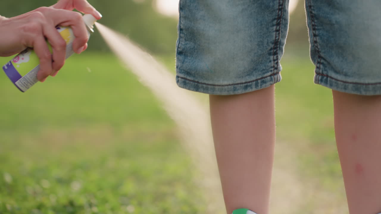 close up of kid leg in denim shorts being sprayed with insect repellent mist in sunny park, hand pressing nozzle, protective moment during outdoor family outing in green grass setting
