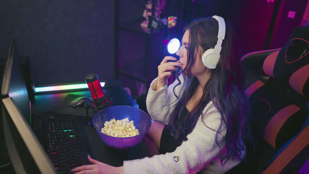 Young gamer woman wearing headphones grabs popcorn from bowl, dusts hand on cozy cardigan then continues typing keyboard in vibrant neon lit studio while microphone