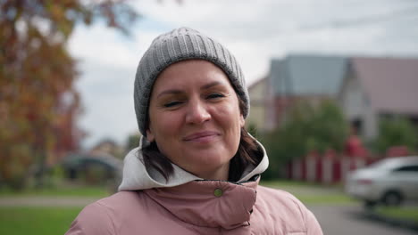 middle-aged woman in a pink jacket and grey beanie smiles warmly, head tilted slightly, with a blurred background featuring buildings and parked cars