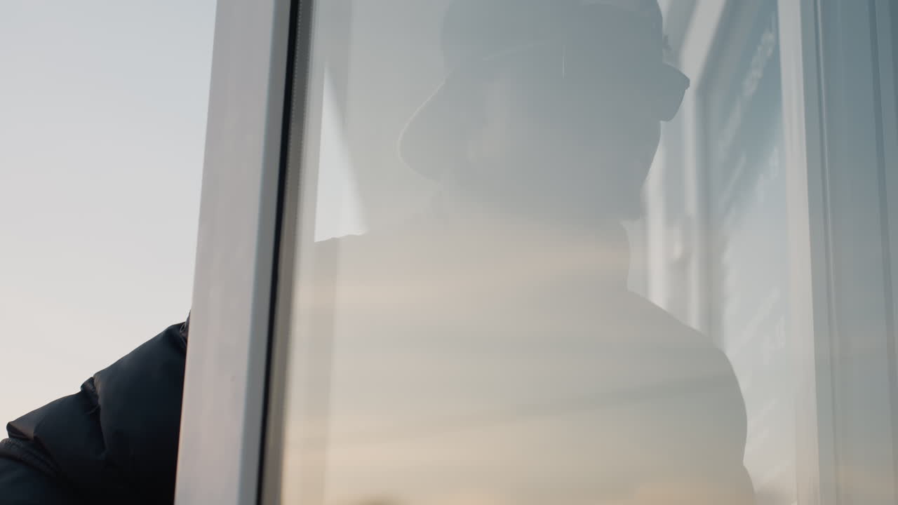 Close up side profile of man wearing black cap and jacket walking outdoors toward mall entrance during winter evening, with soft background of bare trees and pale sky