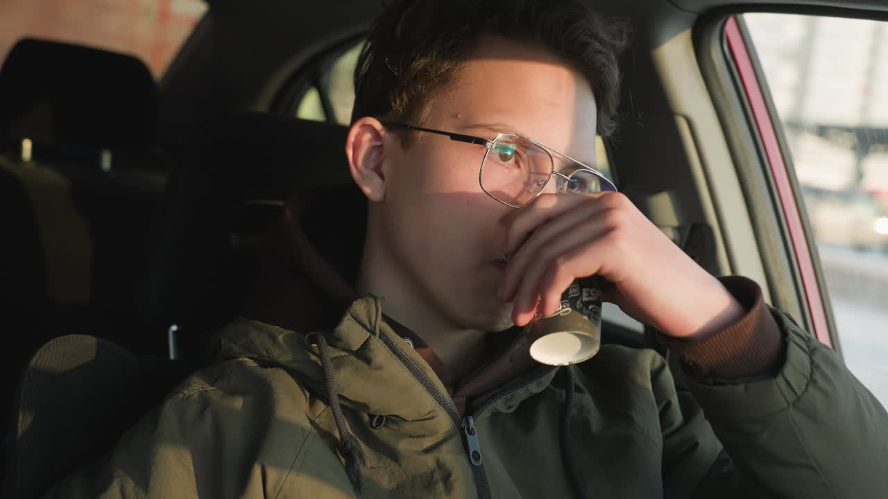 Young boy in glasses and hoodie sits inside parked car sipping coffee from paper cup while holding chocolate candy bar, with warm sunlight casting shadow across his face and car interior