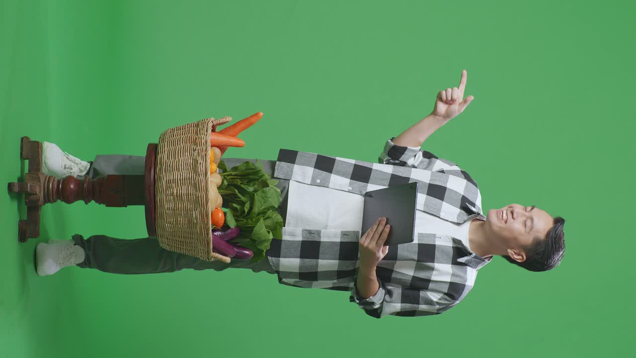 cuerpo lleno de granjero masculino asiático con canasta de verduras usando tableta y apuntando al lado en el fondo de la pantalla verde en el estudio