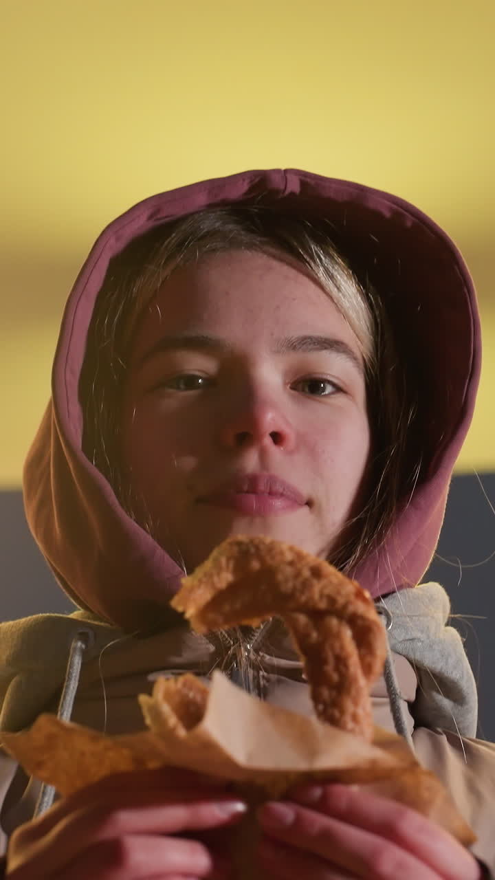 mujer joven de pie al aire libre con la luz de la calle brillante en el fondo, sosteniendo pastel crujiente en la mano, la cálida luz de la ciudad ilumina su cara