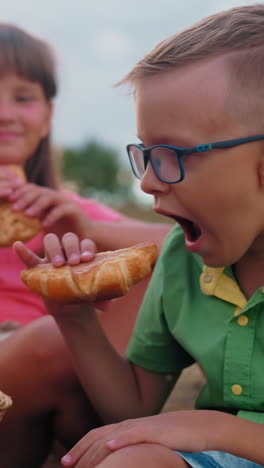 los niños sentados en la hierba disfrutan de pasteles frescos de la canasta de picnic, el niño de camisa verde come inmediatamente mientras que la niña de camisa rosa elige su elección