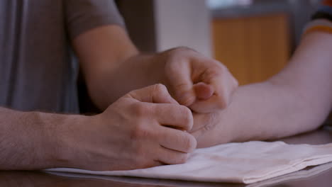 an adult son caregiver assists an elderly mother stroke survivor by gently stimulating and mobilizing her hemiplegic right thumb after stretching her hand; at the kitchen table, on cloth pad.