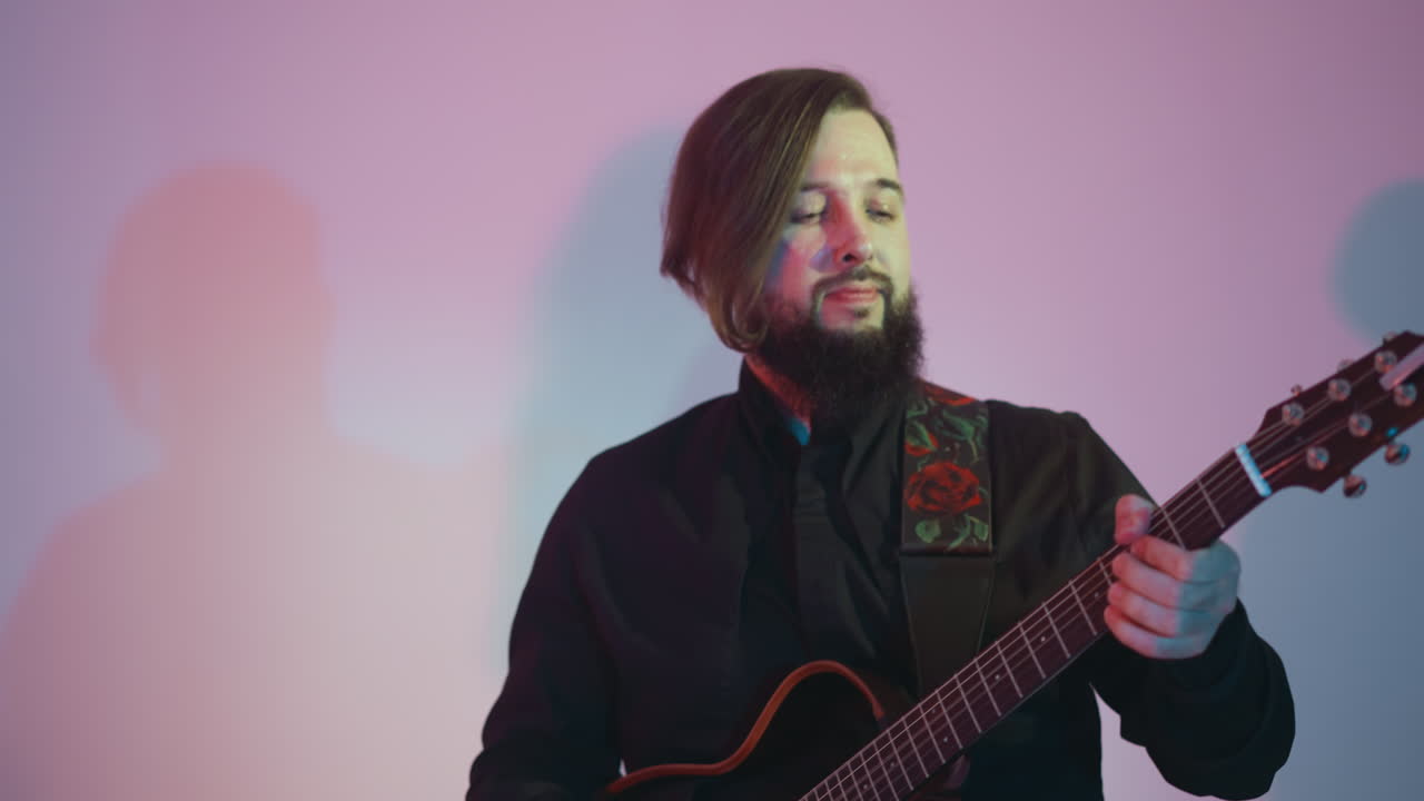 guitarrista barbudo en una camisa negra tocando apasionadamente una guitarra roja, moviendo sutilmente su cabeza contra un fondo blanco con un suave gradiente de luz rosa y azul