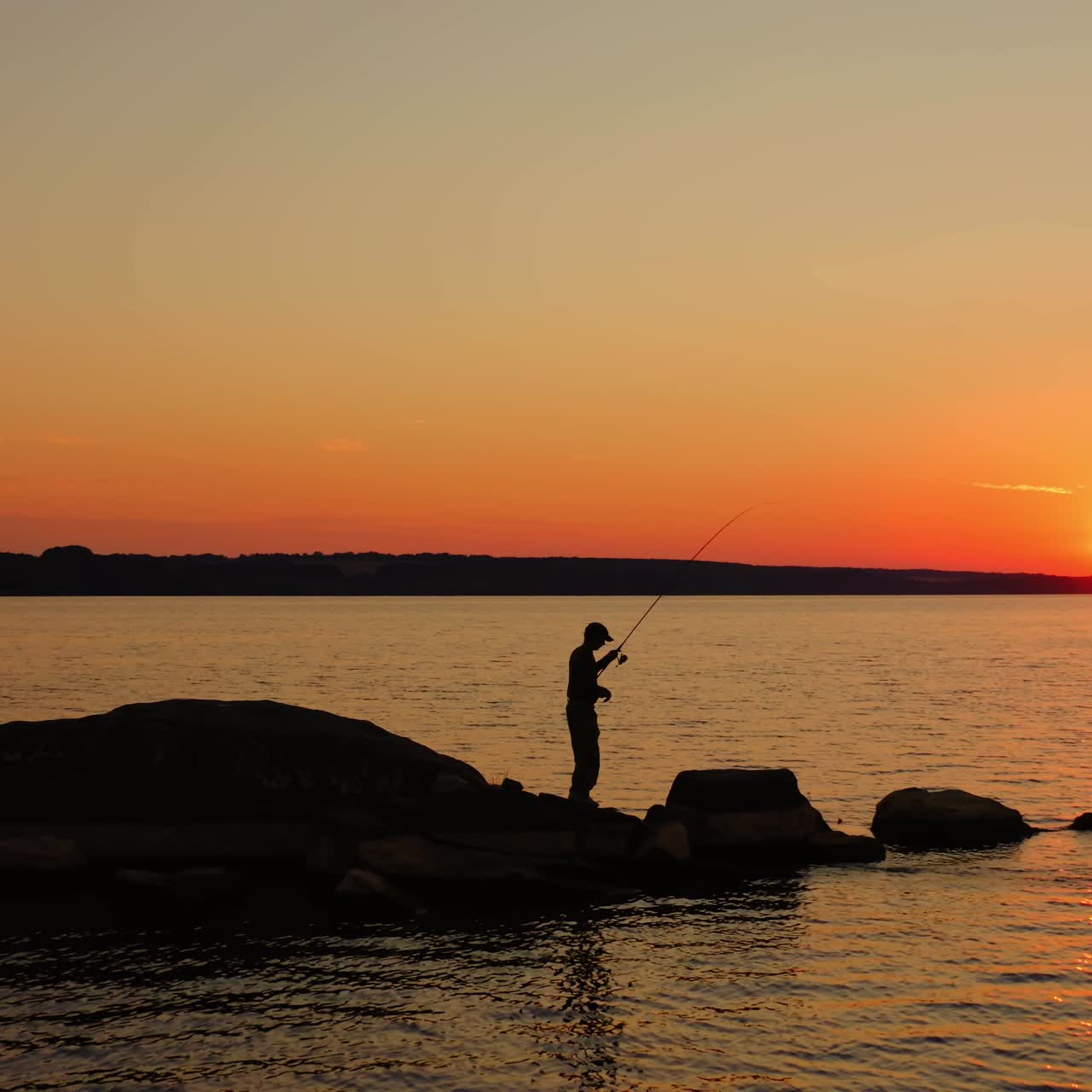 Man fishing at sunset. Silhouette of a man with fishing rod standing on the stone in water in the evening. Fishermen on the lake against orange sunset.