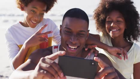 Padre-Afroamericano-Y-Sus-Hijos-Tomándose-Un-Selfie-Con-Un-Teléfono-Inteligente-En-La-Playa