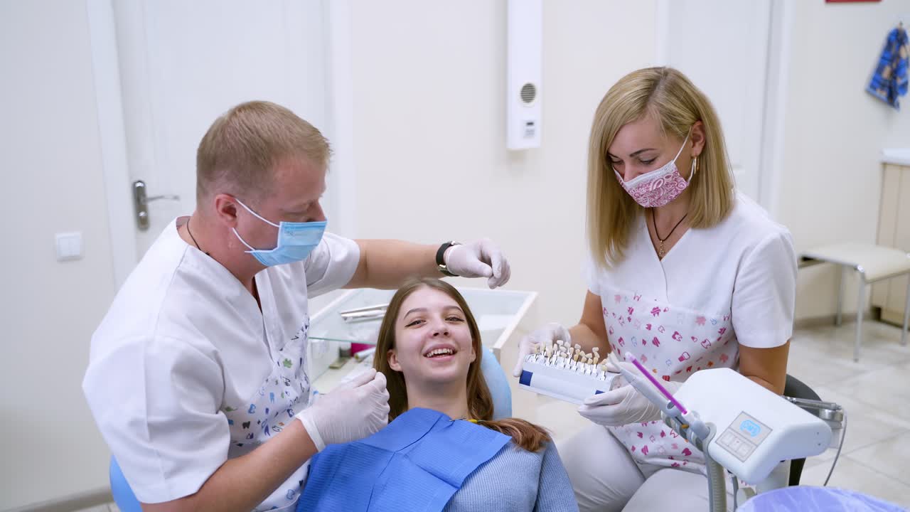 Dentist with assistant work with patient.Professional stomatologist chooses the color of artificial tooth for smiling woman. Benefits of tooth implants.