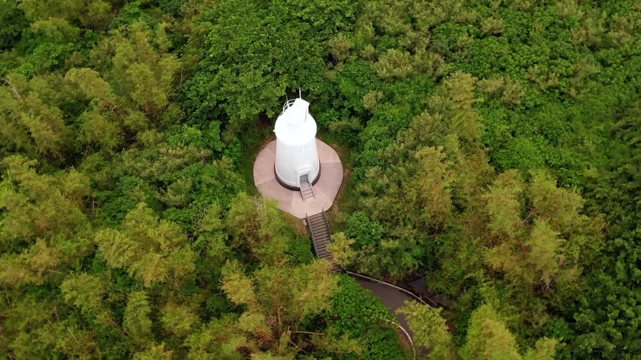 pan circular aéreo, capturando la torre del faro blanco en la cima de la colina que envía rayos de luz para garantizar la seguridad de los barcos en el mar, rodeado de bosque verde en la isla xiaoliuqiu lambai