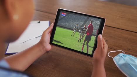 Composite-of-woman-sitting-at-table-with-face-mask,-watching-rugby-match-on-tablet