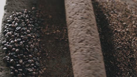 Vista-Superior-En-Cámara-Lenta-De-La-Persona-Que-Muele-Lentamente-Granos-De-Café-Con-Un-Rodillo-De-Roca-En-Una-Piedra-Volcánica