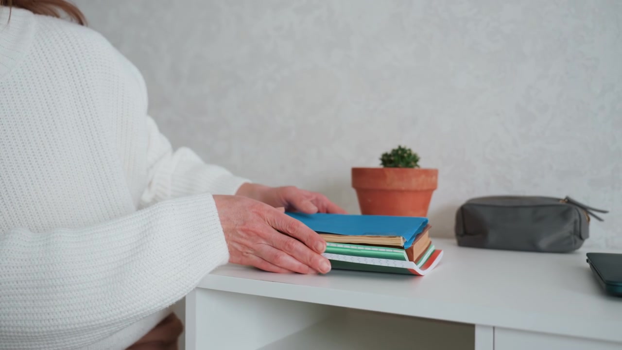 White woman stacking books on desk, wearing cozy sweater beside succulent pot, hands arranging notebooks and journals on tidy workspace, focused preparation for lesson or study routine with minimal
