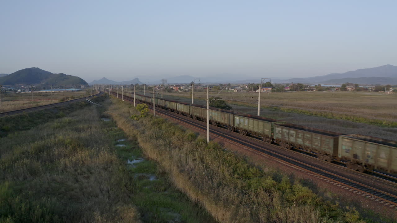 일몰, 러시아 연방을 배경으로 산이 있는 푸른 들판을 통해 긴 빈 석탄 객차 화물을 당기는 기관차
