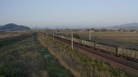 locomotives pulling a long empty coal carriages freight through green fields with mountains in the background on the sunset, russian federation