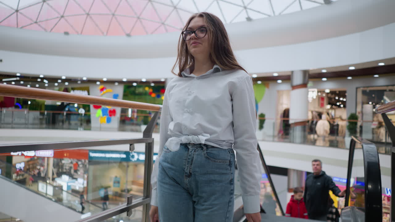 Ung kvinde stiger selvsikkert af en rulletrappe med indkøbsposer. Andre mennesker kan ses bag hende i et livligt, moderne butikscenter