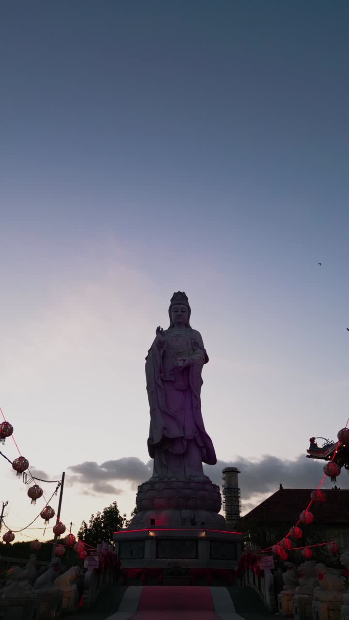 This vertical drone shot highlights a graceful Buddhist statue framed by a traditional temple and the subtle colors of the evening sky emphasizing spiritual harmony and architectural beauty