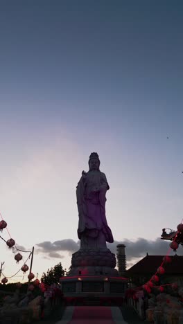 this vertical drone shot highlights a graceful buddhist statue framed by a traditional temple and the subtle colors of the evening sky emphasizing spiritual harmony and architectural beauty