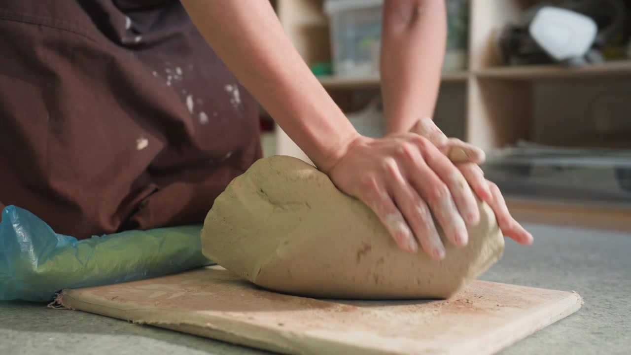Caucasian potter workshop kneading soft clay, hands pressing and folding clay on slab board, apron with clay stains, shelves with tools and jars in warm natural light, tactile texture and focused slow