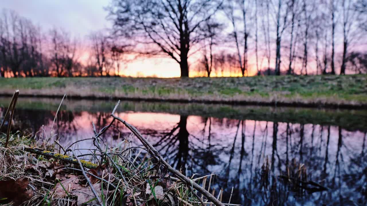 Peaceful stream reflects sunset glow, leafless trees in calm Uzvara Park, Latvia