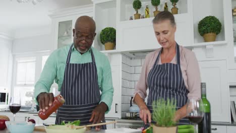 smiling senior diverse couple wearing blue aprons and cooking in kitchen