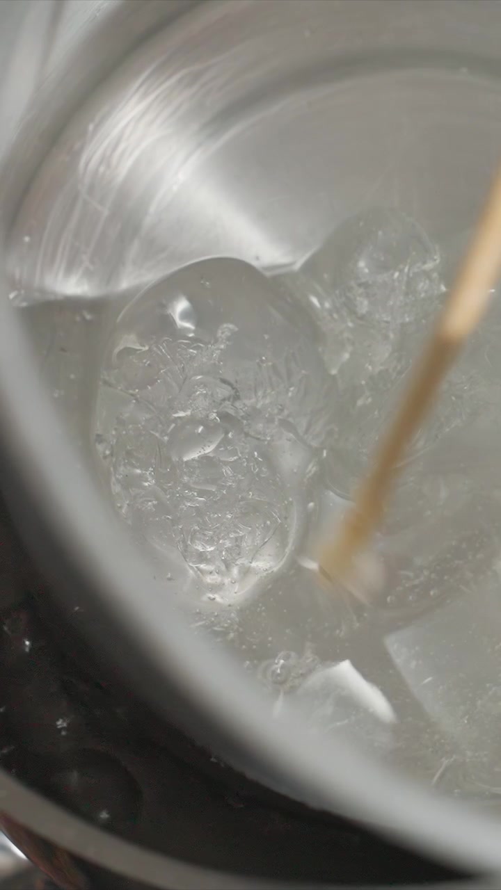 Primo piano di una sostanza trasparente simile a gel mescolata in un contenitore di metallo con un bastoncino di legno. Il gel si addensa e forma bolle visibili mentre viene mescolato, mostrando dettagli della texture sotto luce soffusa.