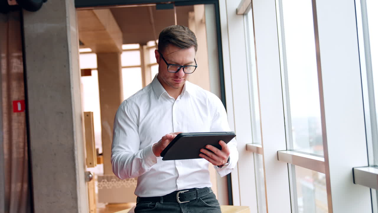 Man is focused on his work on laptop using stylus pencil. Female colleague comes up, puts on glasses and looks at gadget screen.