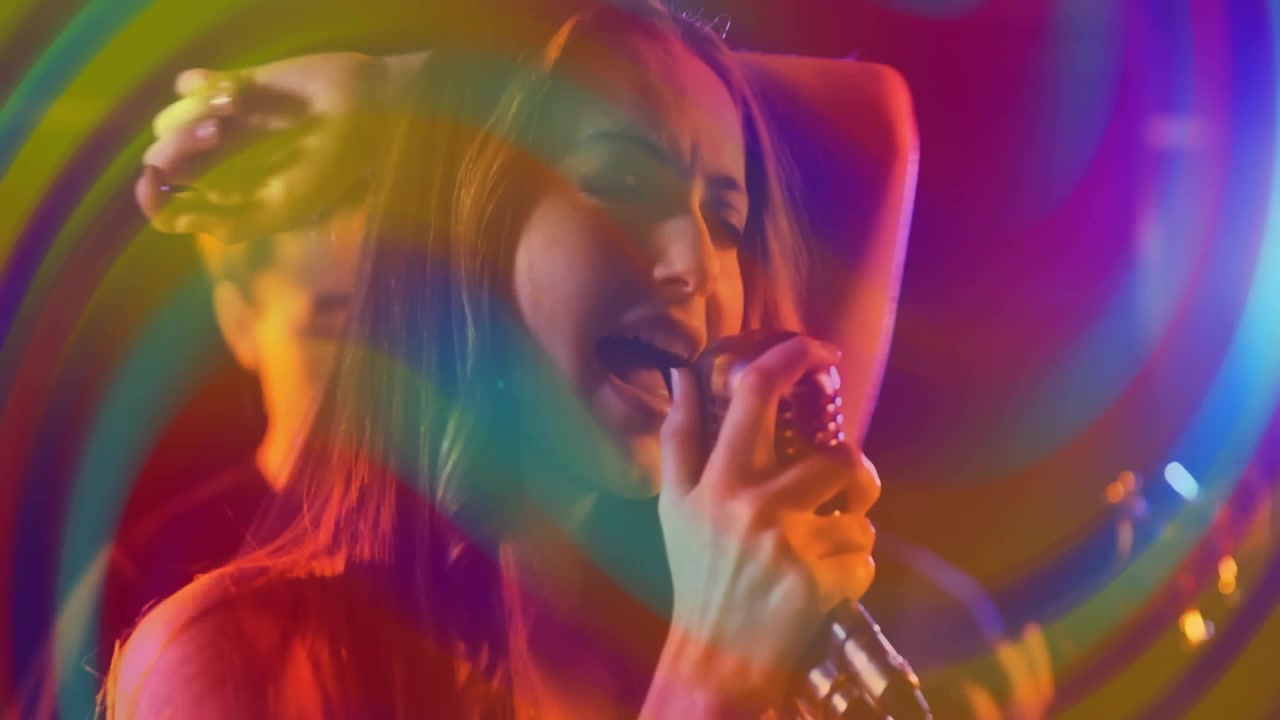 Cantora cantando em microfone retrô enquanto guitarrista toca, com anéis de arco-íris sobrepondo o quadro.