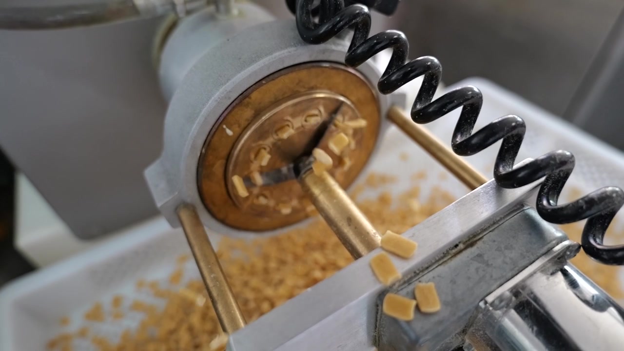 Primo piano dal basso di una taglierina rotante ad alta velocità che taglia pasta appena estrusa in piccoli pezzi prima che cadano in un vassoio di raccolta.