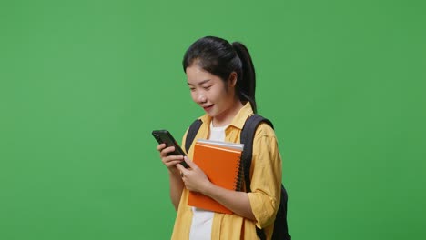 side view of asian woman student with a backpack smiling and using a smartphone while standing in the green screen background studio