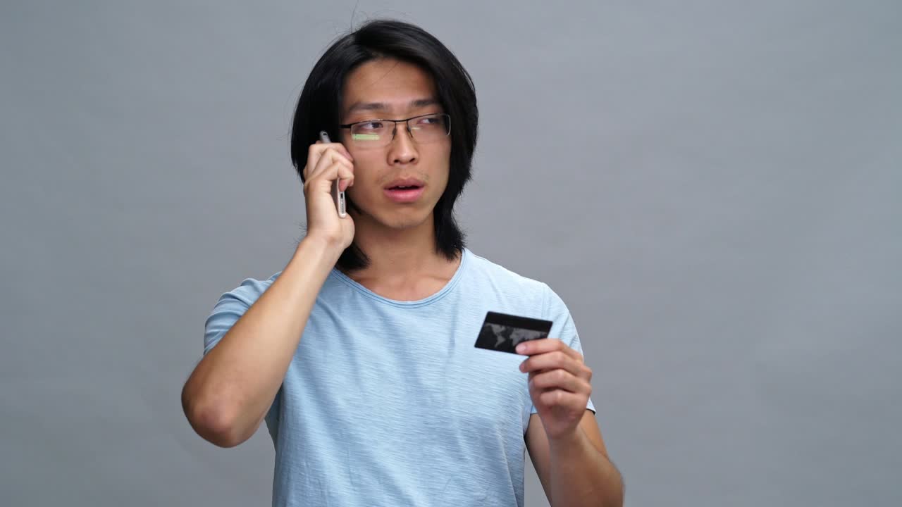Handsome angry young asian man talking by phone while holding credit card