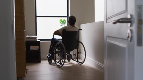 Senior-mixed-race-man-in-wheelchair-wearing-face-mask-and-looking-through-window