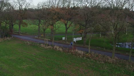 Drohnenaufnahme-Eines-Mannes,-Der-Im-Herbst-Auf-Einer-Kleinen-Straße-Mit-Bäumen-An-Der-Seite-Radelt,-Aus-Der-Luft