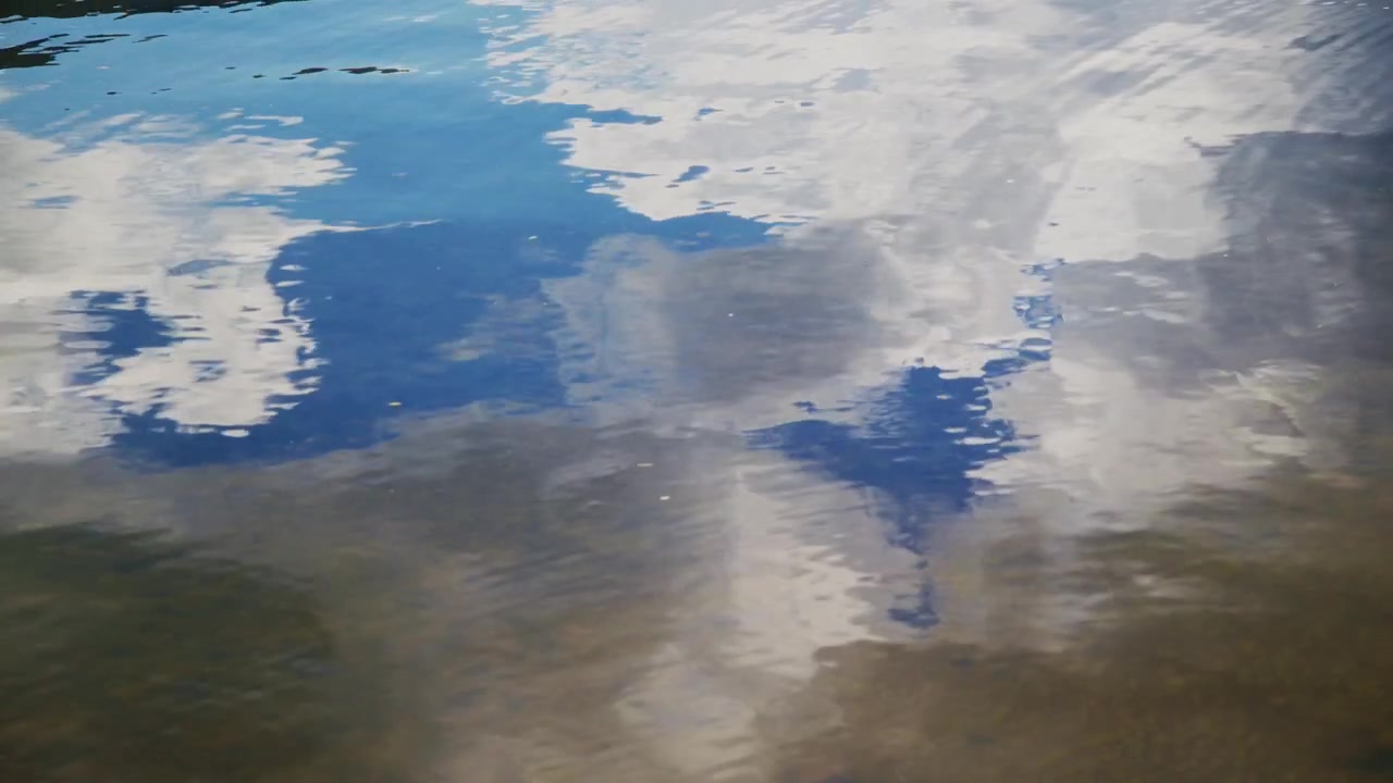Agua tranquila refleja cielo azul y nubes blancas. Suaves ondas distorsionan ligeramente el reflejo. La escena se siente pacífica y serena. Ideal para proyectos de naturaleza, viajes o meditación.